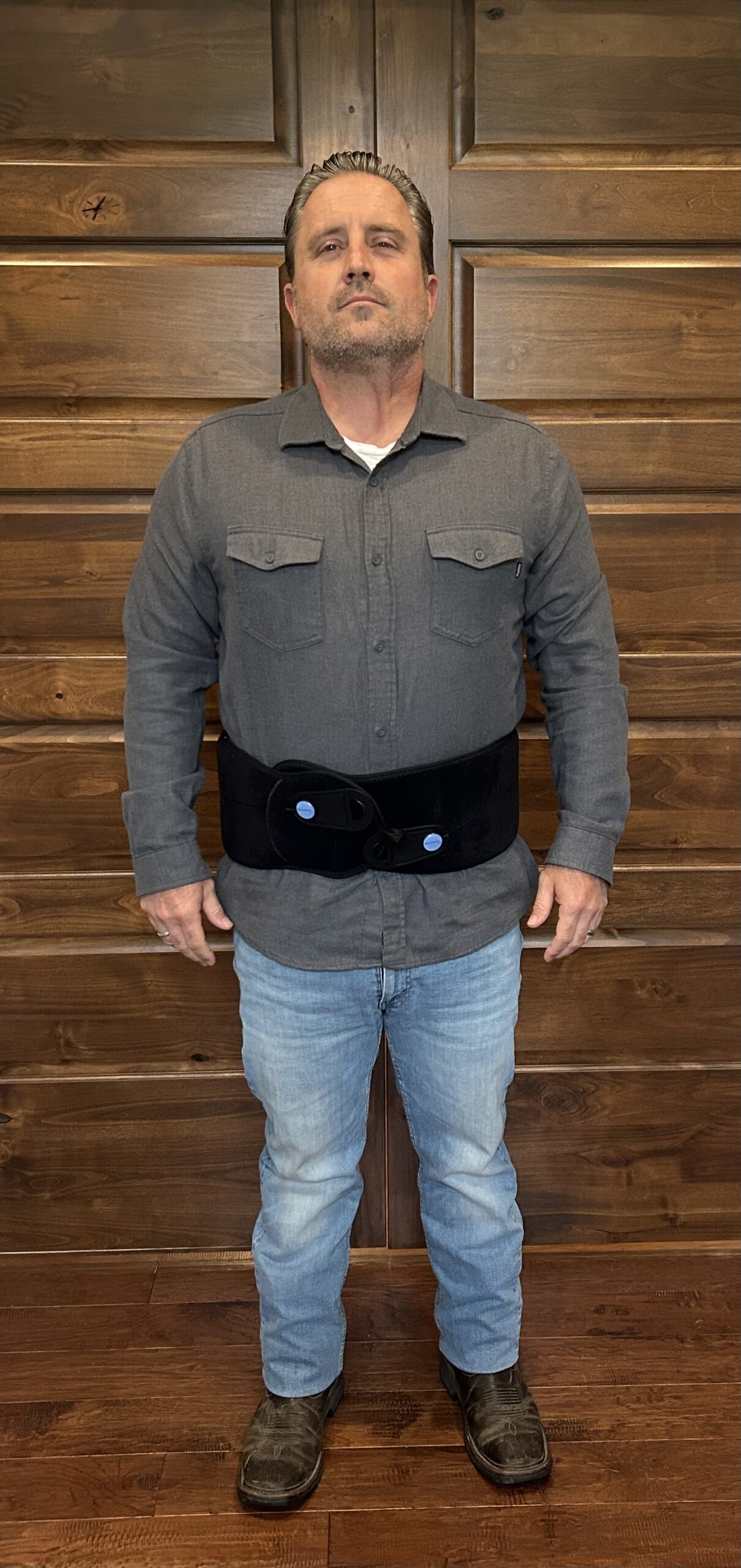 Man wearing a gray button-up shirt and blue jeans, standing against a wooden backdrop, with a supportive black back brace around his waist.