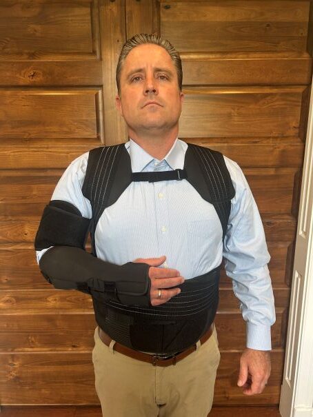 A man in a blue shirt and beige pants wears a black arm brace and torso support, demonstrating medical equipment for rehabilitation.